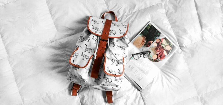 Image of a book bag, open book and sunglasses on a bed with a white comforter. Photo by Cynthia del Río on Unsplash