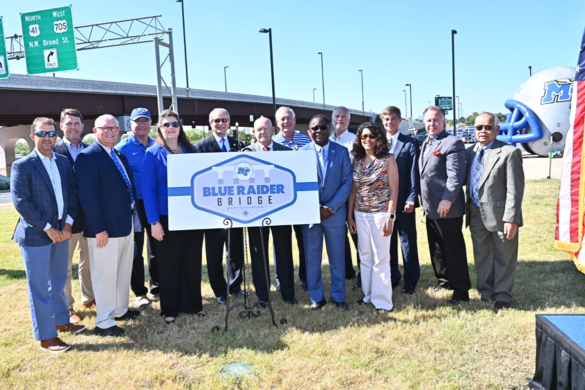 A host of MTSU, Murfreesboro, Rutherford County and state officials stand with a commemorative sign at the Thursday, Sept. 5, dedication ceremony of the newly named Blue Raider Bridge in the background. (MTSU photo by J. Intintoli)