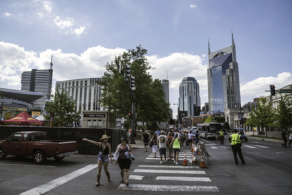 Pedestrians cross Demonbreun Street at Fourth Avenue South in downtown Nashville near the Music City Walk of Fame Park and the Schermerhorn Symphony Center as the city skyline rises behind them.