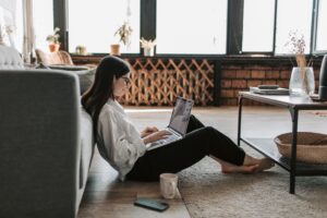A woman sits on the floor in front of her couch, holding a laptop in her lap. 