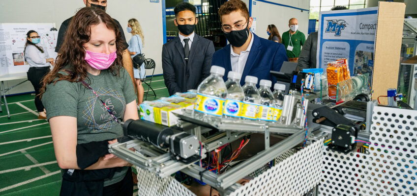 Hannah Davis, left, of Murfreesboro, left, waits for her food and beverage items to drop out of the “Compact Cafeteria” project as team member Jose Rubio explains how the technology works Thursday, April 29, during the annual MTSU Engineering Technology Mech-Tech Expo on the track level inside Murphy Center. The event showcased MTSU seniors’ projects. (MTSU photo by Andy Heidt)
