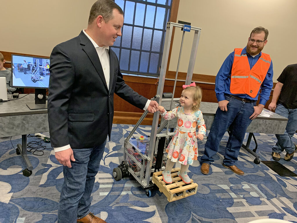 MTSU mechatronics engineering student J.R. Jackson, left, of Lebanon, Tenn., holds 3-year-old daughter Anna Kate’s hand as she rides up and down on the automated forklift his team designed and built for the recent Engineering and Technology Mech-Tech Expo. (MTSU photo by Randy Weiler)
