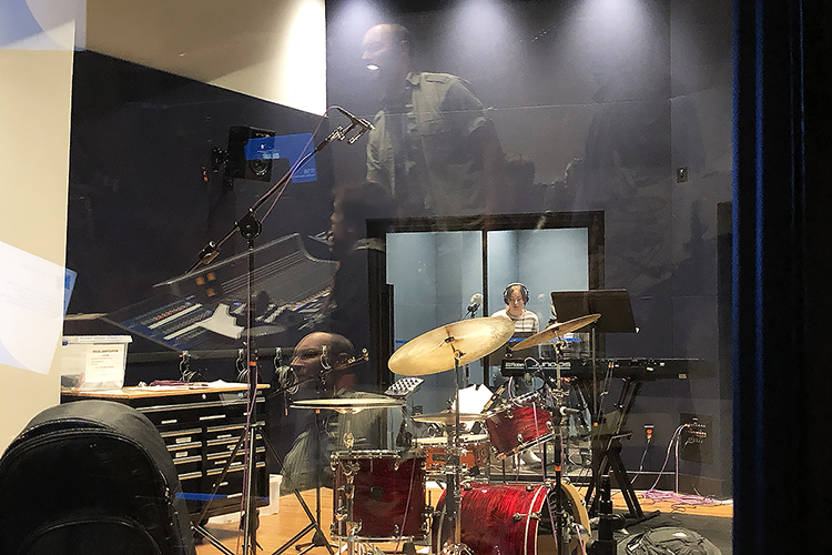MTSU School of Music adjunct professor Matthew Lund is reflected twice in the glass between the control room and recording studio of the Department of Recording Industry’s Studio D in the brand-new Main Street Studios facility during a recent "Studio Saturdays" recording session with students in the Department of Recording Industry Audio Production Program and the School of Music's Commercial Music Ensemble. In the recording booth behind the drum set is sophomore audio production major Alli Wildrick from Allen, Texas, a CME member who provided vocals for some of the day’s tracks. (MTSU photo by Gina E. Fann)