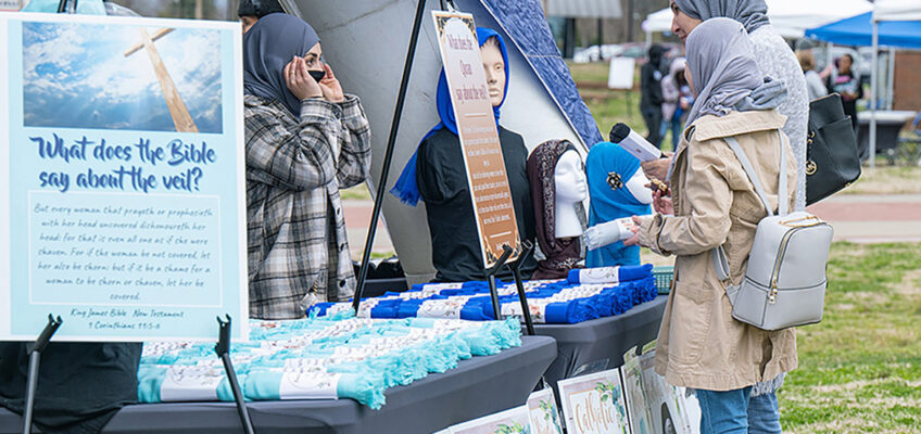 A volunteer explains the hijab, a head covering for Muslim women, to visitors to the Festival of Veils. Posters of various uses of veils in other religions, including Hinduism and Catholicism, are on the grass below tables of free veils. The event was held March 19 on the Student Union Commons. (MTSU photo by Cat Curtis Murphy)