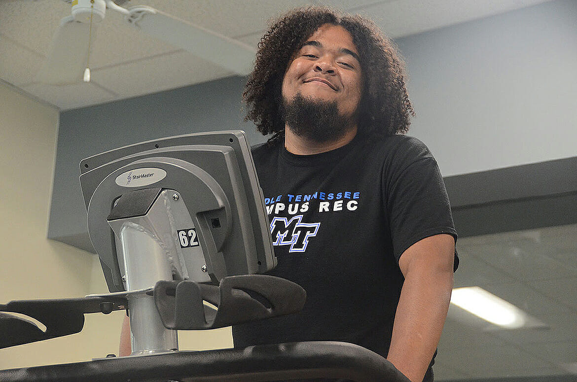 In this June 2018 photo, MTSU audio production major Caleb King of Manchester, Tenn., flashes a smile as he works out on a StairMaster in the cardio room of the MTSU Student Health, Wellness and Recreation Center. King, a supervisor at the Rec, has used the facility toward reaching his 100-pound weight loss goal. (MTSU photo by K. Simpson)