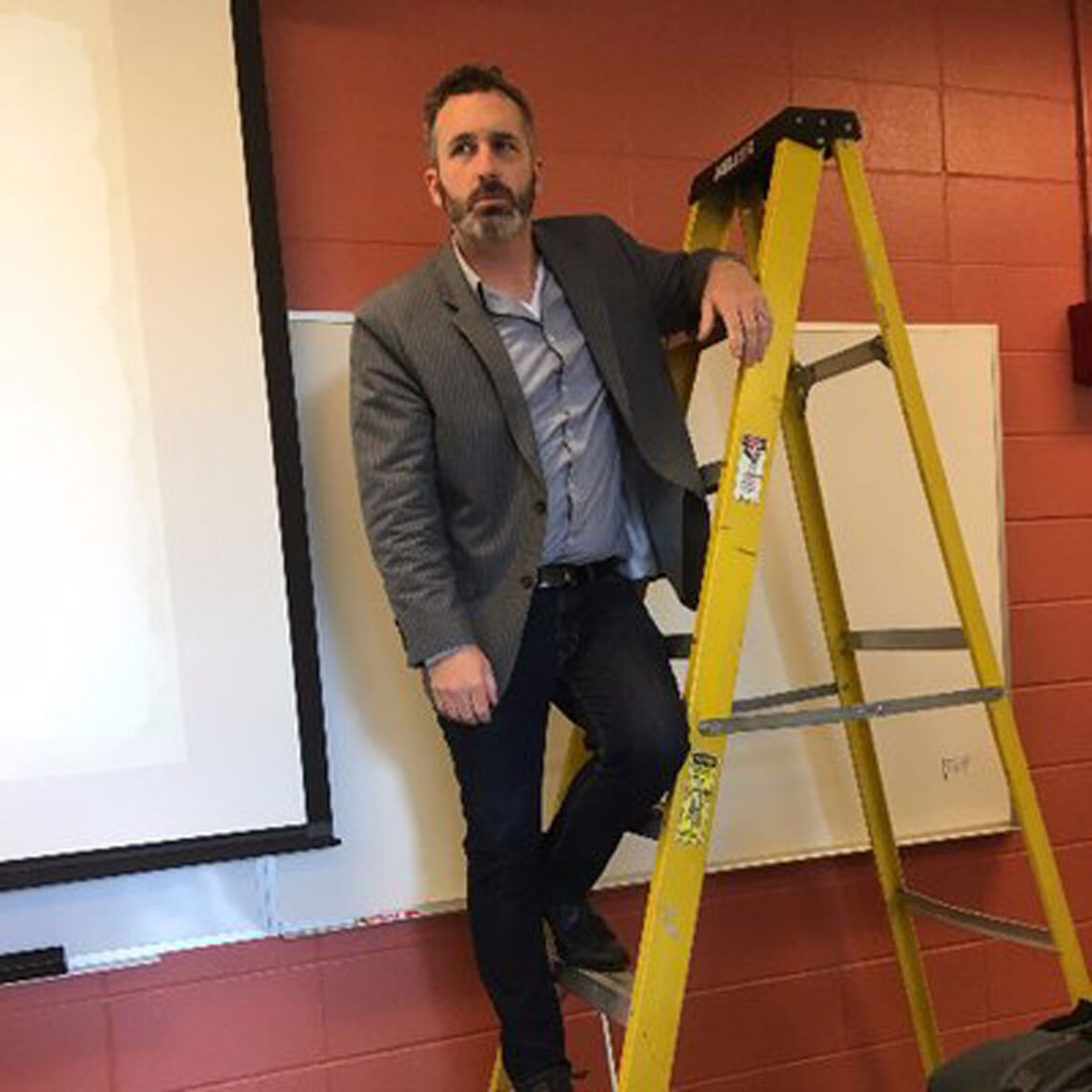 Portrait of Ben Sawyer, professor of History at MTSU, being funny by standing on a ladder in a classroom.