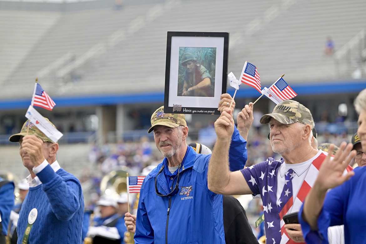 Middle Tennessee State University alumnus and U.S. Army Veteran Bob Lamb (Classes of 1969 and ’77), center, of Murfreesboro, Tenn., walks across Horace Jones Field in Floyd Stadium in Murfreesboro carrying a framed photo of his longtime friend and veteran, Bud Morris (’69, ’75), in November 2024, during the 42nd annual MTSU Salute to Veterans and Armed Forces game during the halftime parade by service branches to theme songs performed by the Band of Blue. Morris is expected to rejoin his military brothers this year during the special halftime ceremony on Saturday, Nov. 8. (MTSU file photo by Andy Heidt)