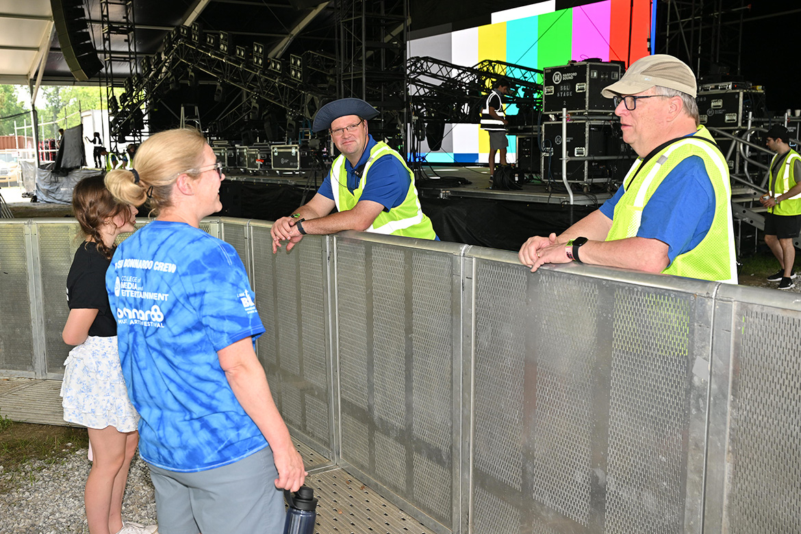 Robert Gordon, right, interim chair of the Department of Media Arts at Middle Tennessee State University in Murfreesboro, Tenn., chats with Katie Foss, front left, director of MTSU’s School of Journalism and Strategic Media, as students set up on stage in the background Wednesday, June 11, at the 2025 Bonnaroo Music and Arts Festival in Manchester, Tenn. Looking on, center, is Mike Forbes, director of MTSU Technical Systems and an adjunct media arts lecturer. (MTSU photo by James Cessna)