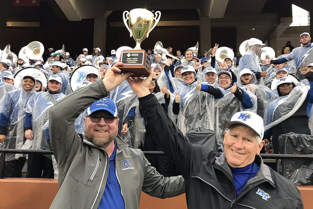 Ray Wiley, left, associate director of MTSU Campus Recreation and a longtime American Red Cross volunteer, joins MTSU Director of Athletics Chris Massaro to hoist the annual “blood battle” challenge trophy aloft Saturday, Nov. 30, at at the MTSU vs. Western Kentucky University football game in Bowling Green. Cheering behind them are members of the MTSU Band of Blue. MTSU won the annual blood drive contest for the third straight year and the seventh time since it began in 2010. (Photo submitted)