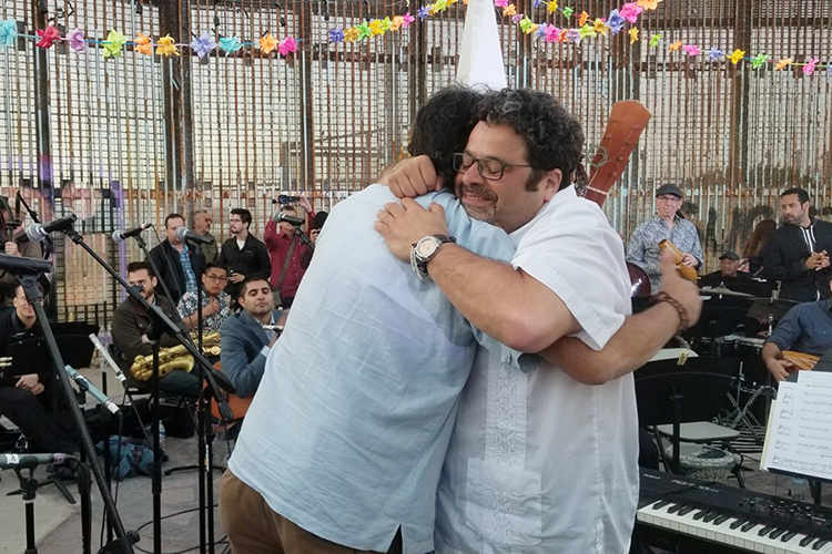 Sunlight shines through the barrier as musicians Arturo O'Farrill, left, and Jorge Francisco Castillo hug after performing at the border wall in Tijuana, Mexico, in this May 2018 file image provided by makers of the documentary “Fandango at The Wall.” (file image courtesy of FandangoWall.com)