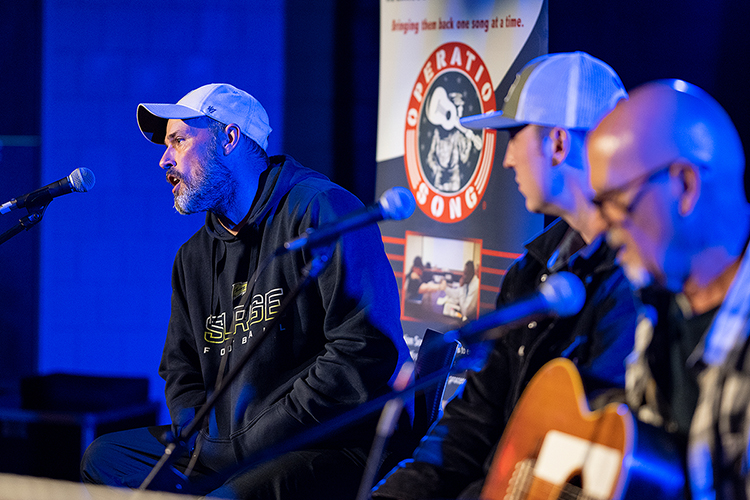MTSU student veteran Jason Mettert of Murfreesboro, left, a senior human geography major, offers the audience some background information about "Jump 64," a song composed from discussing his military service during the annual Operation Song writers' retreat at the university. Looking on before they perform the song in MTSU's Keathley University Center Theater Oct. 28 are his co-writers, senior commercial songwriting major Patrick "PC" Strickland from Hoover, Alabama, and Grammy nominee Billy Montana, who’s written for Jo Dee Messina, Sara Evans and Garth Brooks and helped guide the gentle twists of Mettert’s story. MTSU's Charlie and Hazel Daniels Veterans and Military Family Center and the Commercial Songwriting Program have hosted the event since 2016. (MTSU photo by Cat Curtis Murphy)
