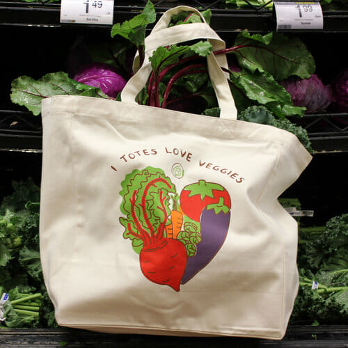 A reusable grocery bag, illustrated with vegetables, hangs in front of shelves of vegetables. 
