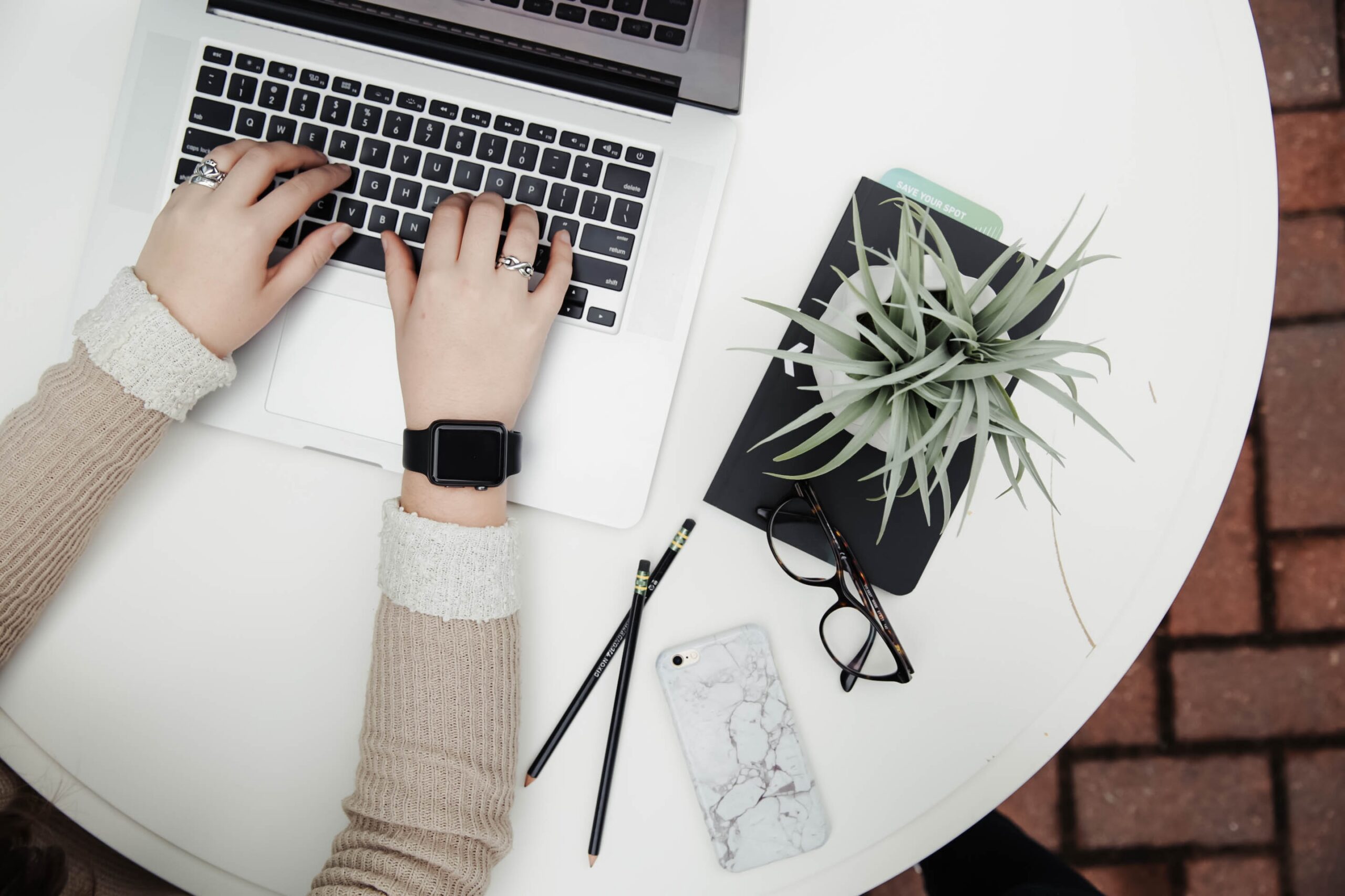 Hands typing on a laptop. Table has a plant, cell phone, eyeglasses, pencils, and a plant.
