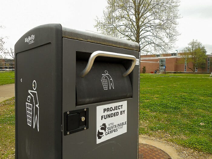 An MTSU recycling bin sits on a sidewalk, surrounded by green grass and a tree.